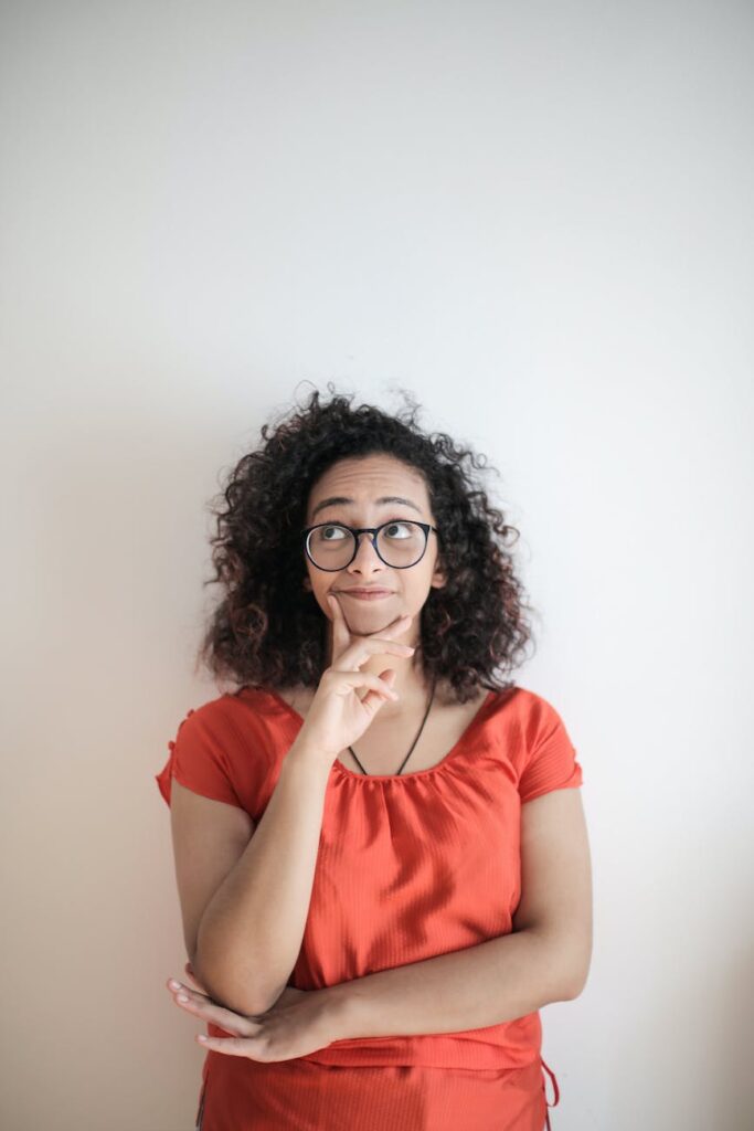 portrait photo of woman in red top wearing black framed eyeglasses standing in front of white background thinking